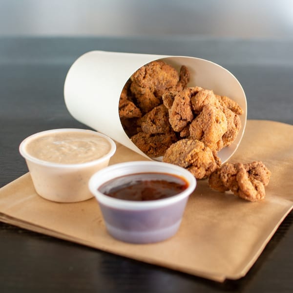 Crispy fried chicken bites spilling from a white cup, with two dipping sauces on a brown paper.
