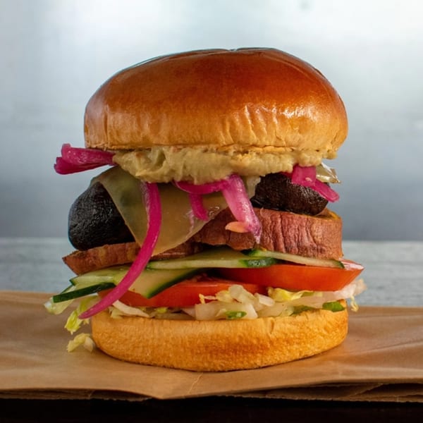 A tall Buns Burgers burger with a thick patty, avocado, pickled onions, and fresh vegetables.
