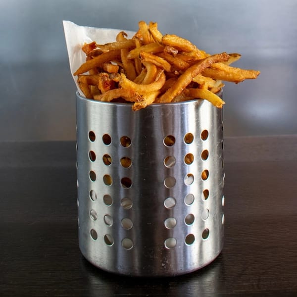 Crispy golden french fries, served in a perforated metal holder with white paper, on a dark table.